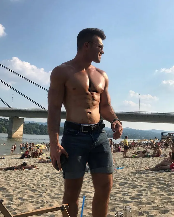 Homme torse nu, debout sur une plage, portant un short en jean et des lunettes de soleil