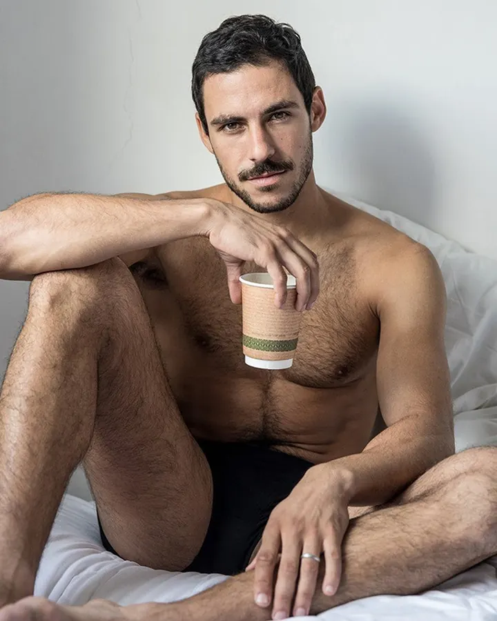 Homme torse nu, assis sur un lit blanc, tenant un gobelet en papier