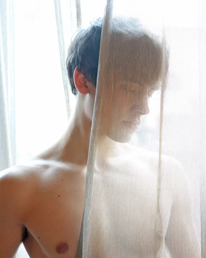 Homme torse nu, cheveux bouclés, portant plusieurs colliers de coquillages, regard perdu dans le lointain, extérieur.