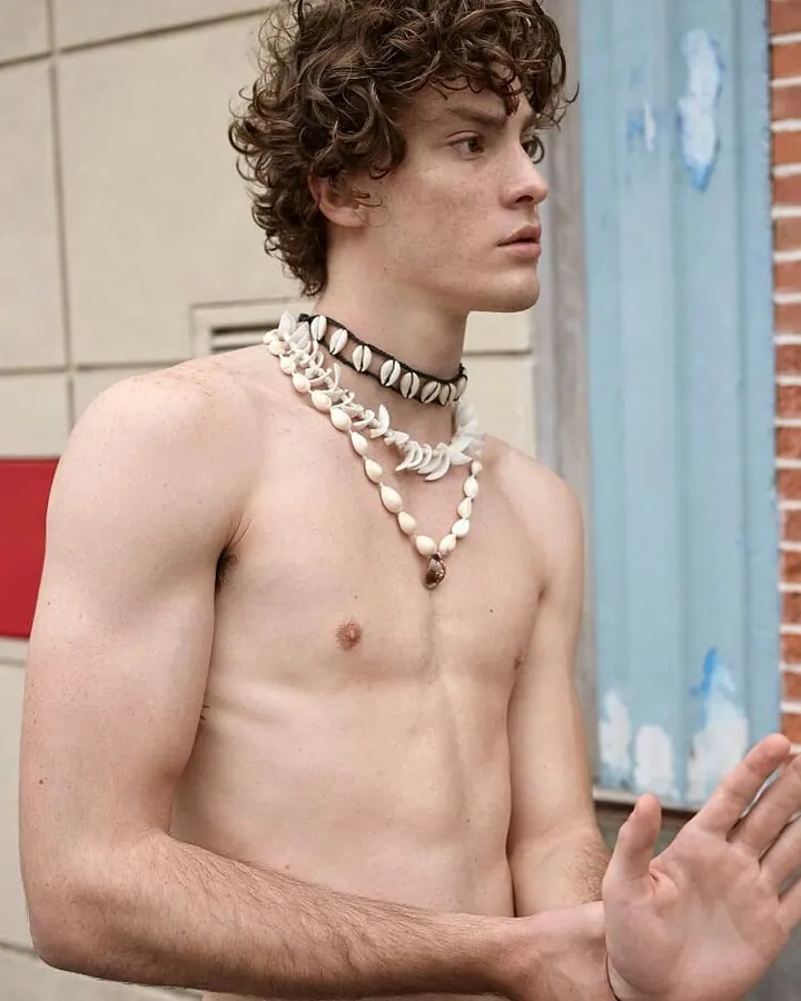 Homme torse nu, peau foncée, tatouage sur le torse, portant un boxer blanc Calvin Klein, debout dans une pièce, regard confiant.