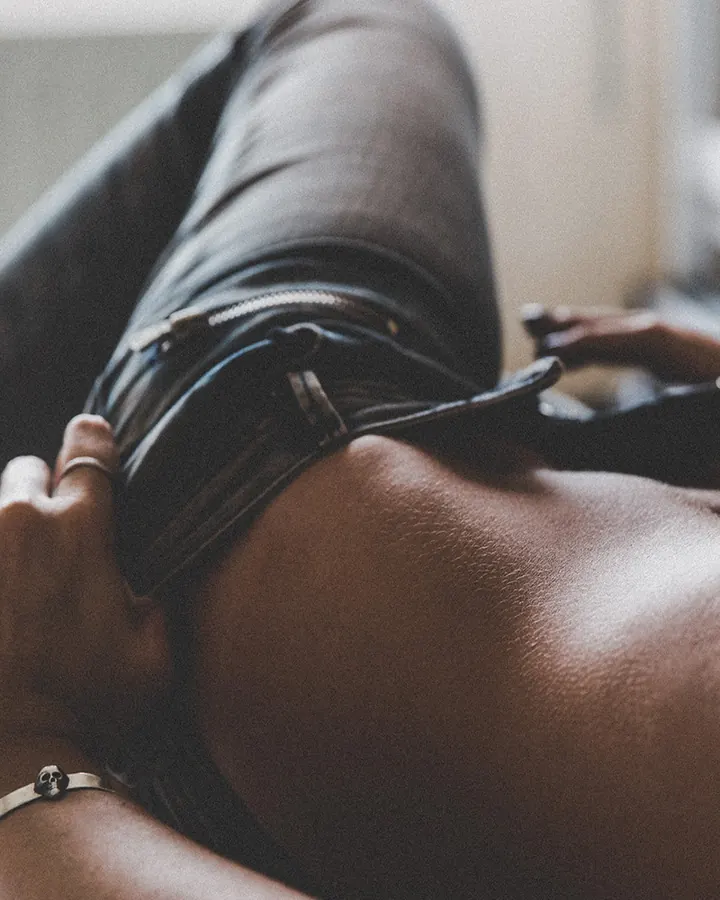 Homme noir allongé, torse nu, main tenant le zip de son pantalon en cuir noir, bracelet crâne à la main, lumière douce.