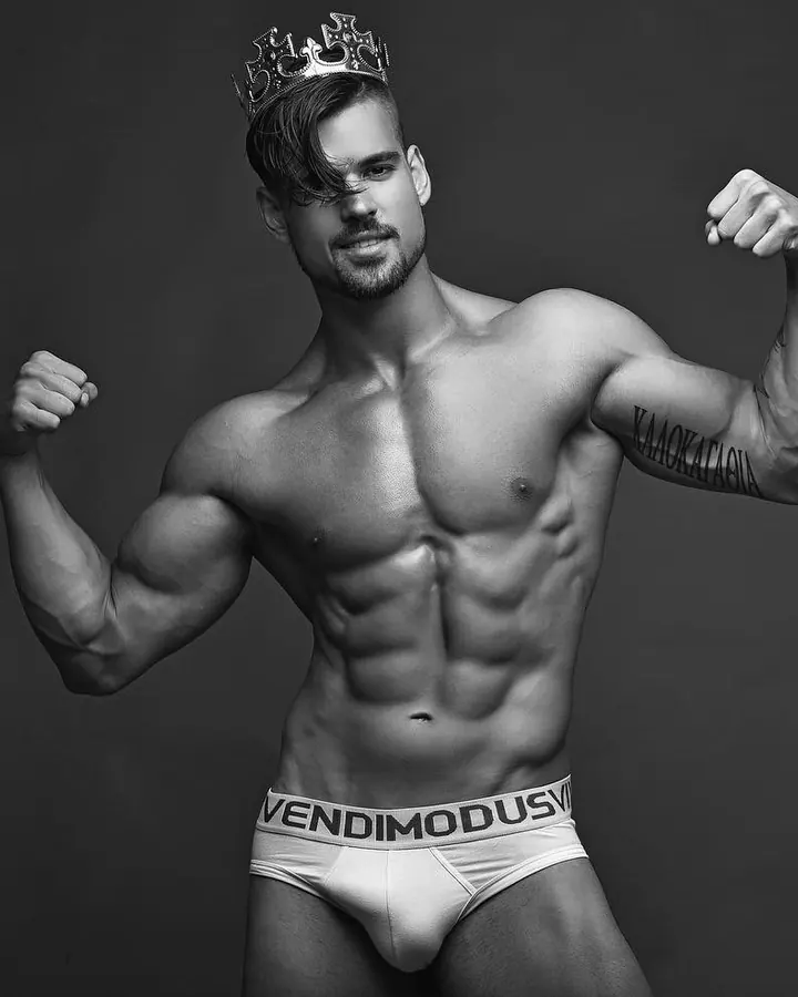 Homme torse nu, cheveux foncés, regard intense, assis sur un lit, couvert par une couette blanche, photo en noir et blanc.