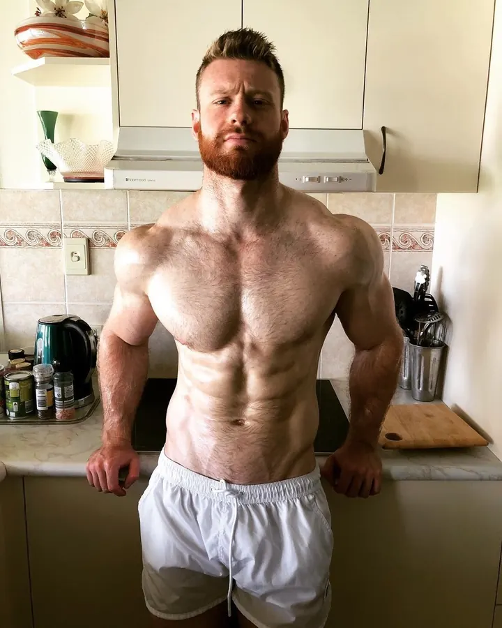 Homme torse nu, barbe rousse, poitrine poilue, debout dans une cuisine, mains sur le plan de travail, regard sérieux.