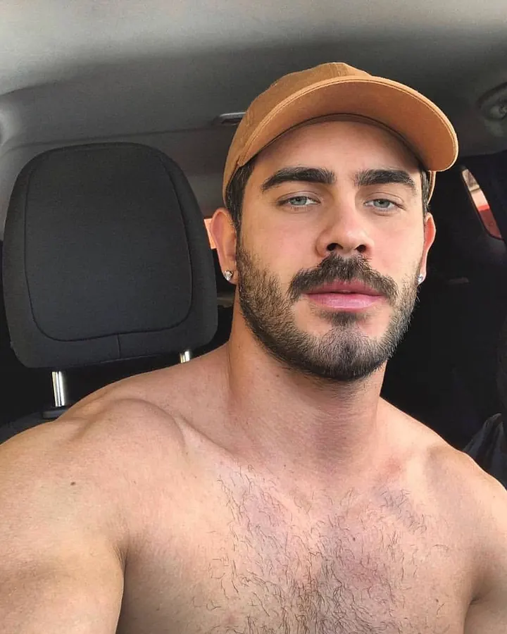 Homme torse nu dans une voiture, portant une casquette marron, regardant directement l'appareil