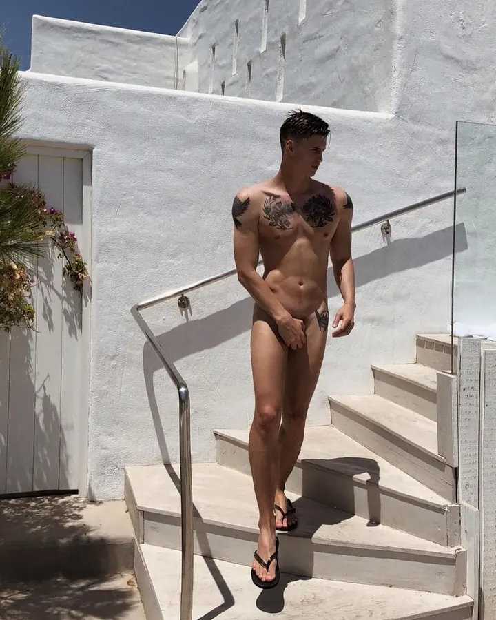 Homme torse nu sur une terrasse, portant un slip blanc à motif flamants roses, une casquette rose et des lunettes de soleil