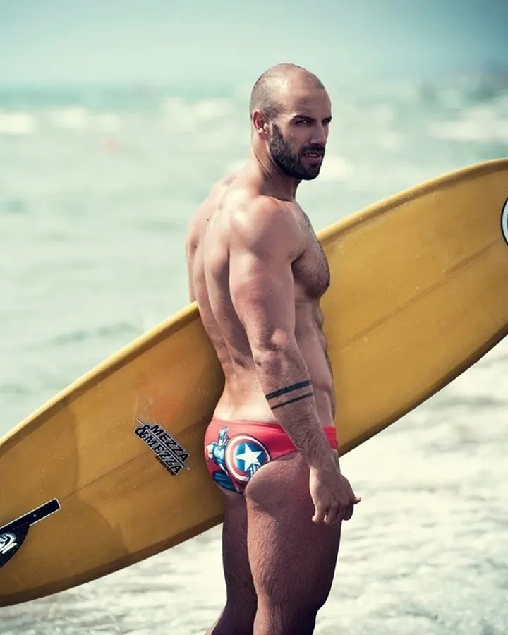 Homme torse nu sur une plage, portant un maillot de bain rouge Captain America, tenant une planche de surf