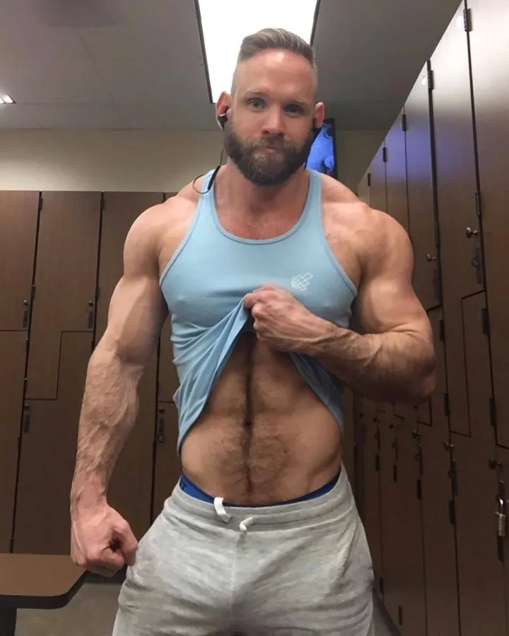 Homme torse nu, en gilet bleu, dans un vestiaire, tenant son haut ouvert, regardant l'appareil photo.