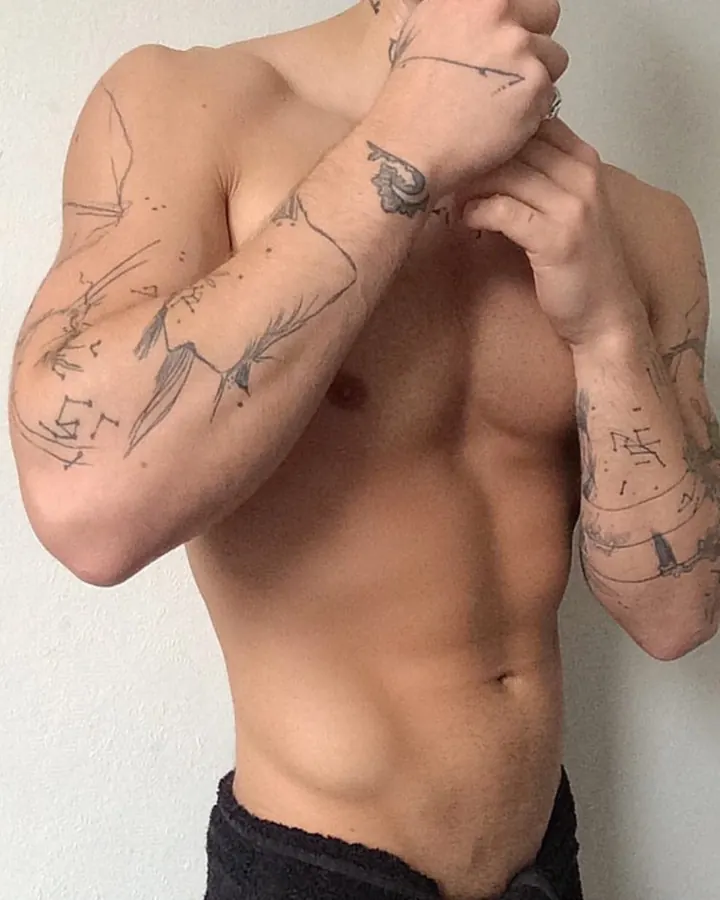 Homme torse nu, bras tatoués, mains croisées devant la poitrine, devant un mur blanc