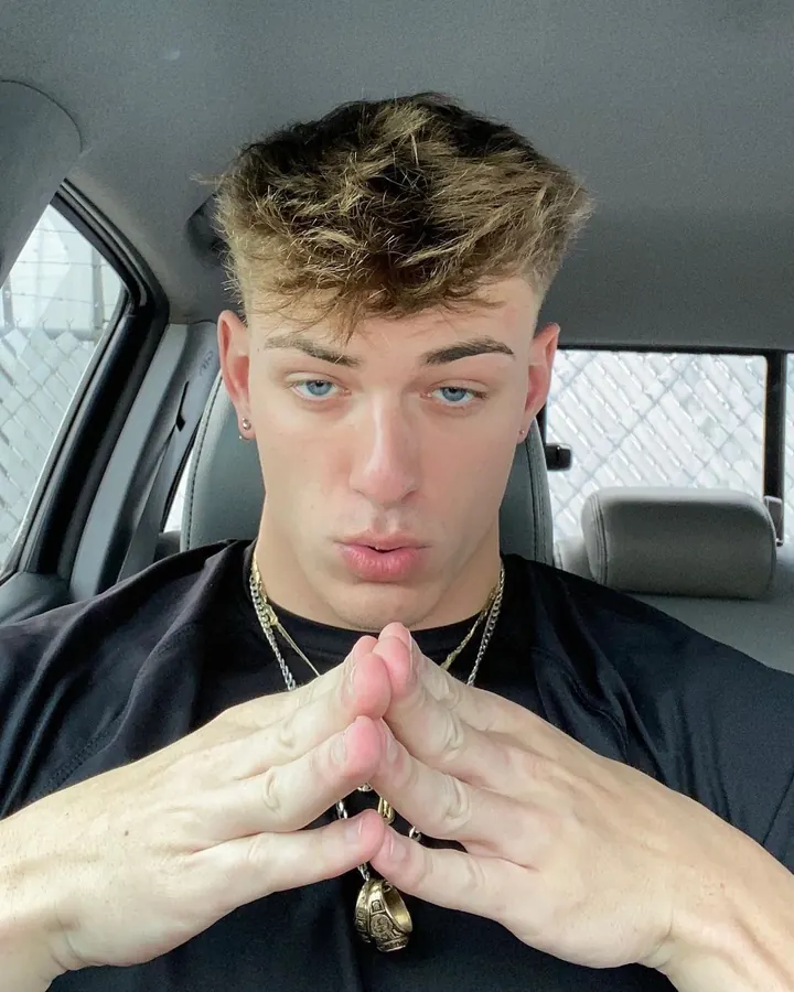 Jeune homme torse nu, portant un t-shirt noir, assis dans une voiture, mains jointes