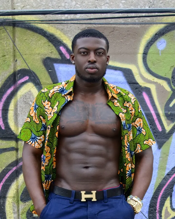 Homme torse nu, en chemise colorée ouverte, devant un mur tagué, regardant l'appareil photo avec assurance.