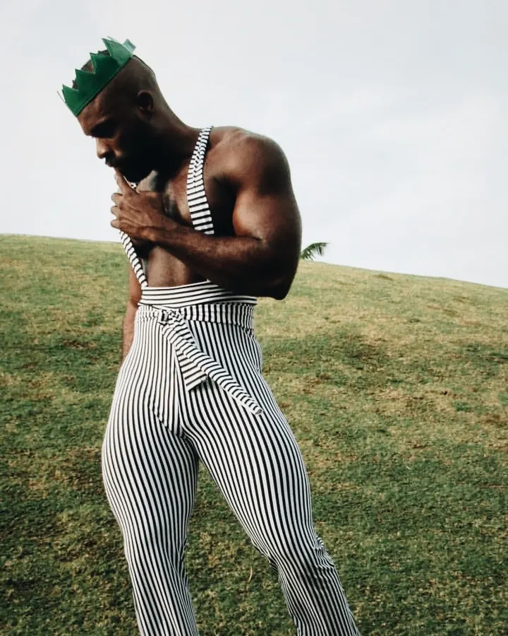 Homme noir en chemise noire, portant des lunettes de soleil, une couronne verte, un haut rayé noir et blanc, un pantalon rayé noir et blanc, les mains sur le torse, debout sur une colline herbeuse.
