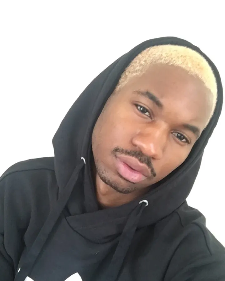 Homme noir en sweat à capuche noir, avec une frange blonde, regardant directement la caméra, sur fond blanc.