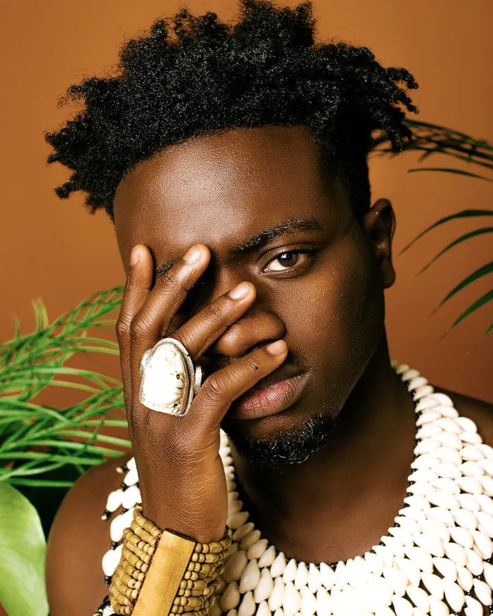 Homme noir en tenue traditionnelle africaine avec collier de coquillages, portant une grande bague blanche, couvert de bracelets, la main sur le front, devant un fond orange avec des plantes vertes.