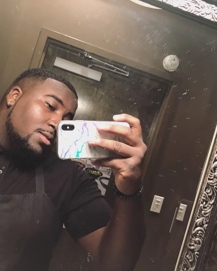 Homme noir prenant un selfie dans un miroir, portant un polo noir, un tablier noir, avec une barbe courte, un piercing à l'oreille, tenant un téléphone blanc avec un motif coloré.