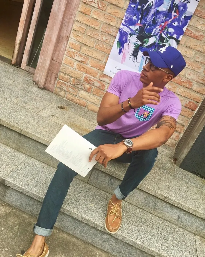 Homme noir assis sur des marches en pierre devant un mur de briques, portant un t-shirt violet, un jean bleu, une casquette bleue et des chaussures marron, tenant un papier blanc.