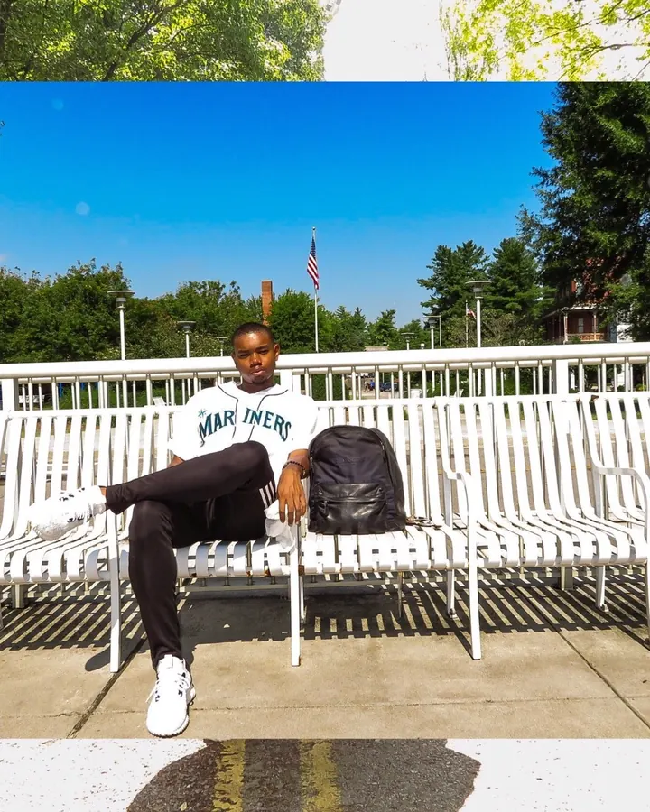 Homme noir assis sur un banc blanc en plein jour, portant un maillot blanc et un pantalon noir, avec un sac à dos noir à côté de lui.