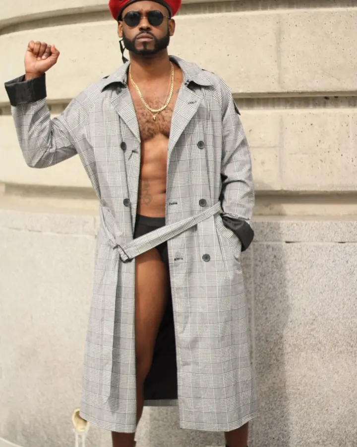 Homme noir portant un long manteau gris à carreaux, un bonnet rouge, des lunettes de soleil, un collier doré, torse nu, sous-vêtements noirs, bottes noires, levant le poing, devant un mur de pierre.