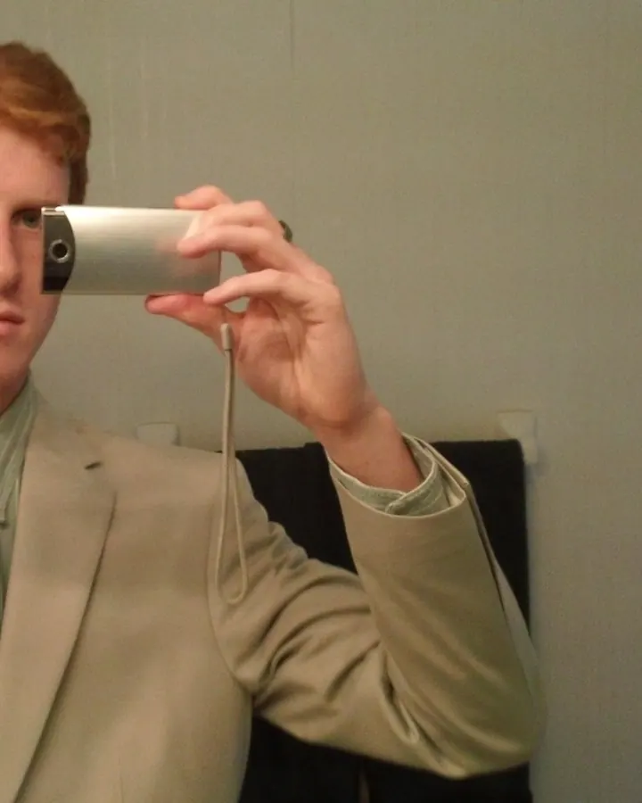 Homme en veste noire, assis dans une voiture, regardant l'appareil photo avec une expression neutre.