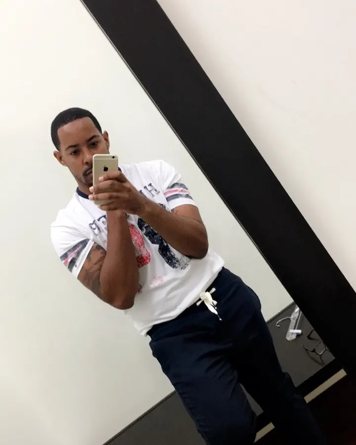 Homme aux cheveux courts noirs, en t-shirt blanc et pantalon bleu, prenant un selfie dans un miroir.