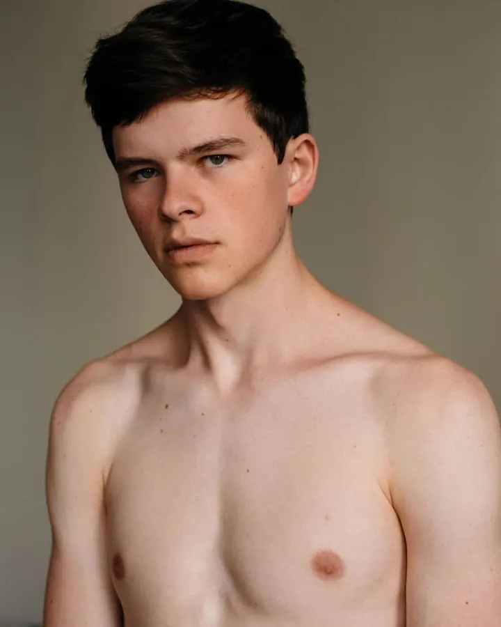 Jeune homme aux cheveux bruns, torse nu, regardant directement l'appareil photo, fond neutre.
