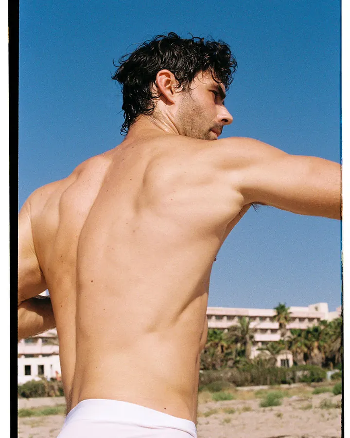 Homme aux cheveux foncés bouclés, torse nu, dos visible, en extérieur sous un ciel bleu.