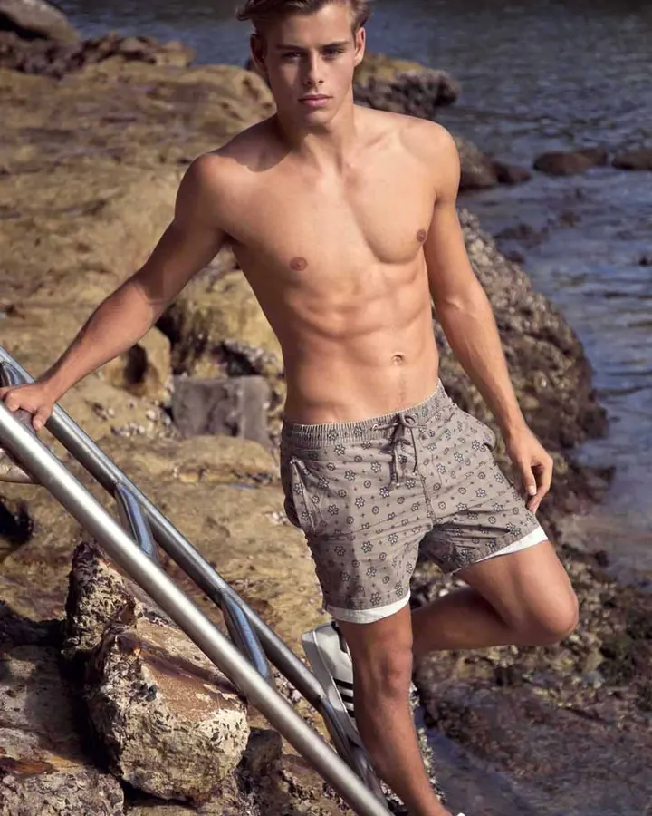 Jeune homme torse nu, cheveux blonds, posant sur des rochers près de l'eau, portant des shorts imprimés.