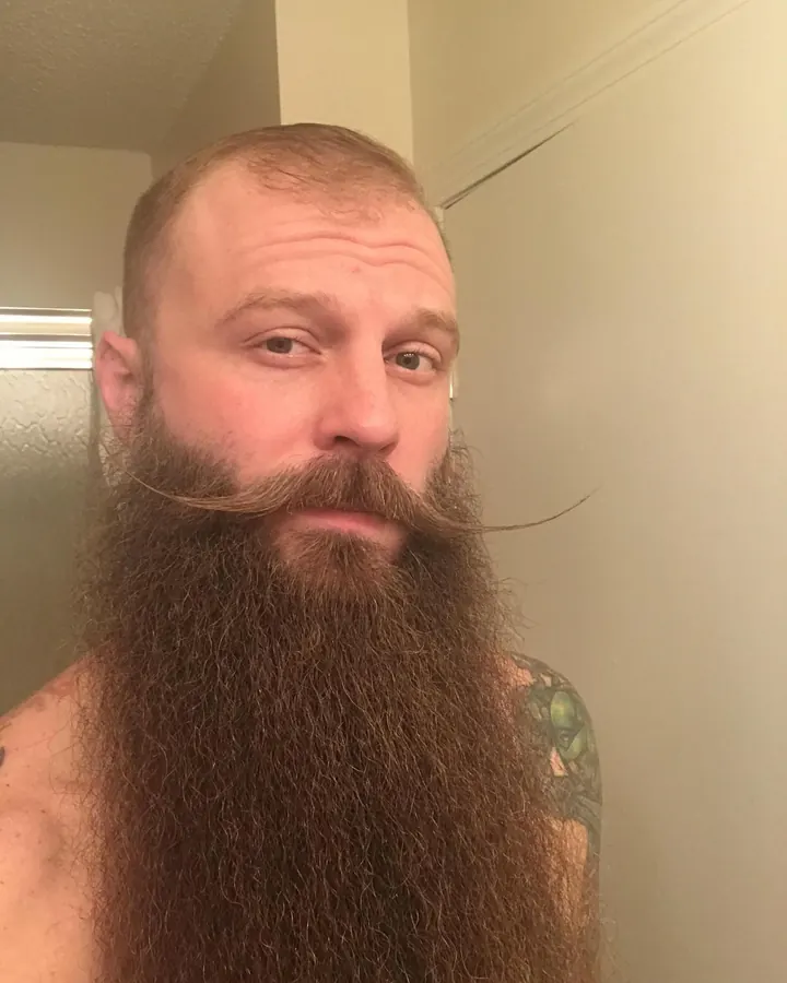 Homme barbu, cheveux ras, regardant l'appareil photo dans une pièce intérieure.