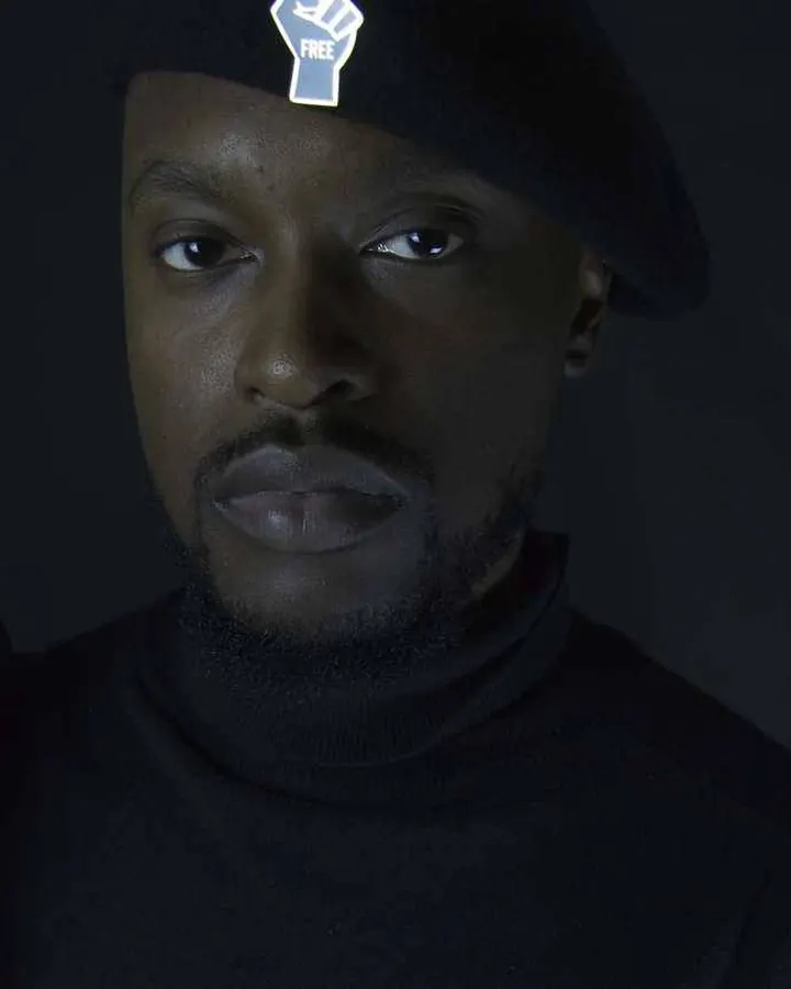 Homme noir portant un bonnet noir avec logo lumineux, vêtu de noir, regardant directement l'appareil photo.