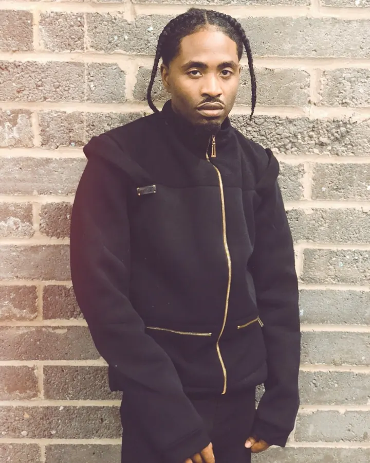 Homme noir portant une veste noire avec fermeture dorée, debout devant un mur de briques.