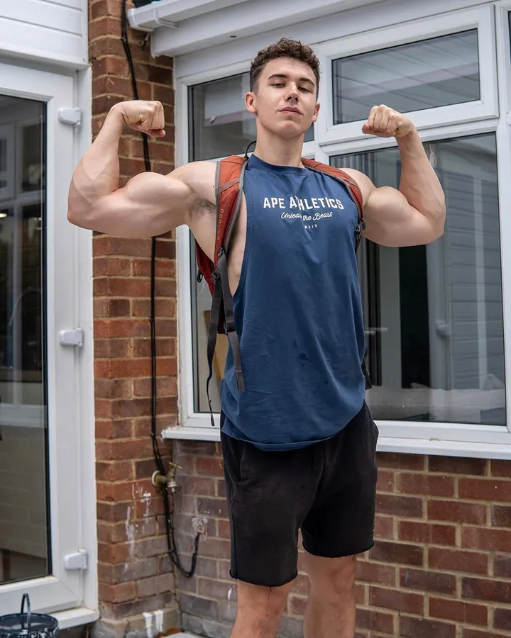Jeune homme musclé portant un débardeur bleu, faisant des flexions devant une maison en briques.