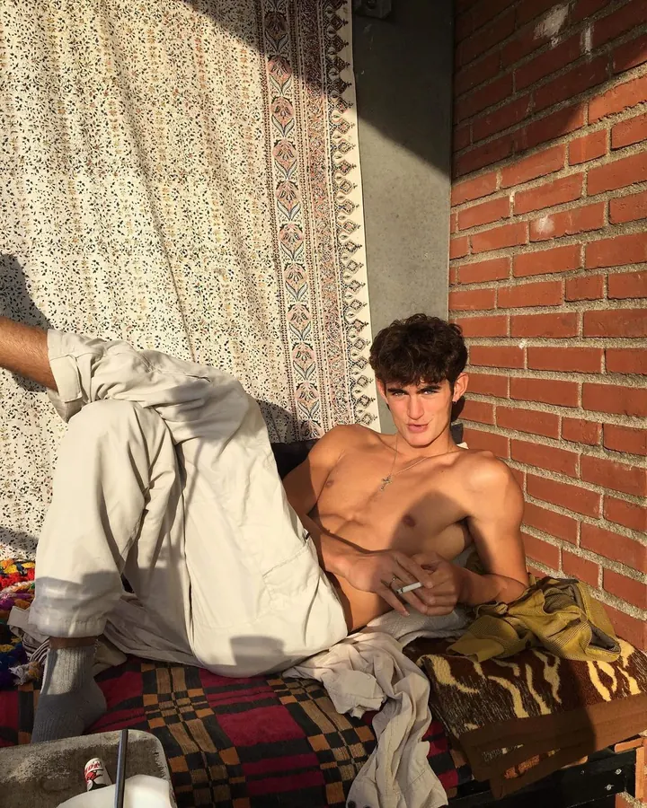 Jeune homme torse nu, allongé sur un lit, fumant une cigarette, entouré de couvertures et de tissus colorés.