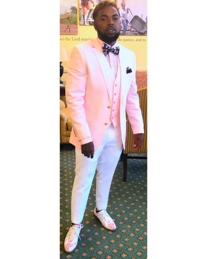 Homme noir portant un costume rose pâle avec nœud papillon, debout dans une pièce.