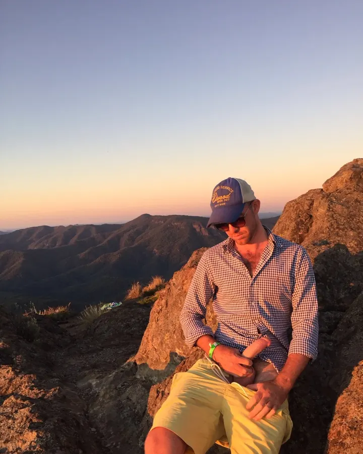 Homme barbu, portant un chapeau de baseball, assis sur un rocher, regardant vers le bas, paysage montagneux en arrière-plan.