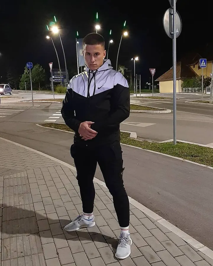 Jeune homme en tenue de sport noir et blanc, debout sur un trottoir la nuit, les mains croisées devant lui.