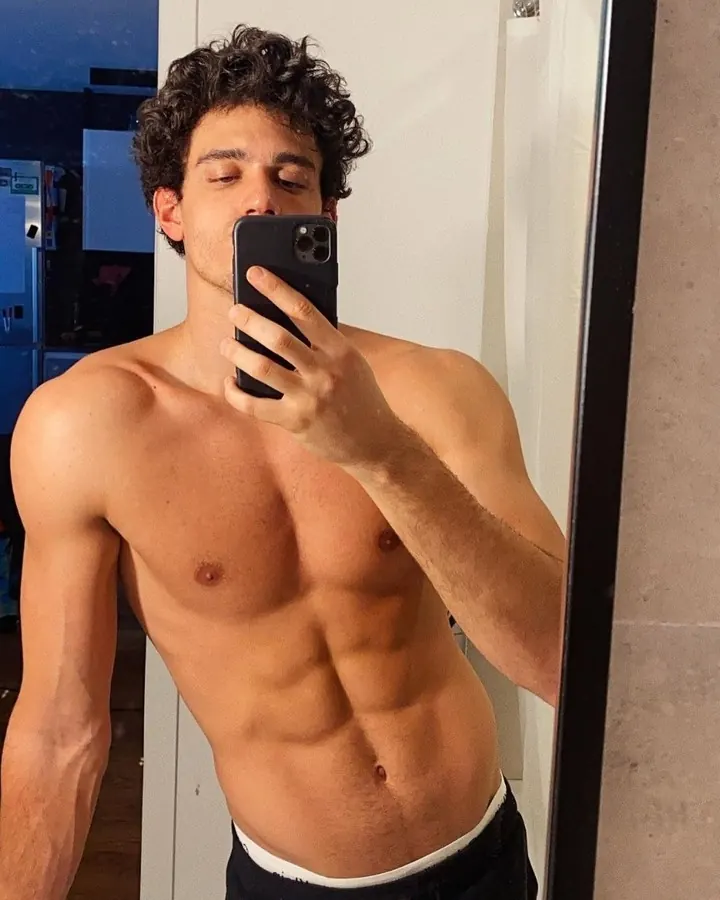 Jeune homme torse nu prenant un selfie devant un miroir, cheveux bouclés, regard fermé.