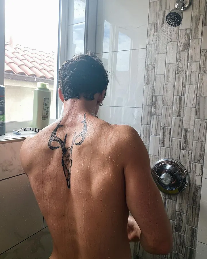 Homme torse nu, tatouage sur le dos, sous la douche, regardant vers le bas.