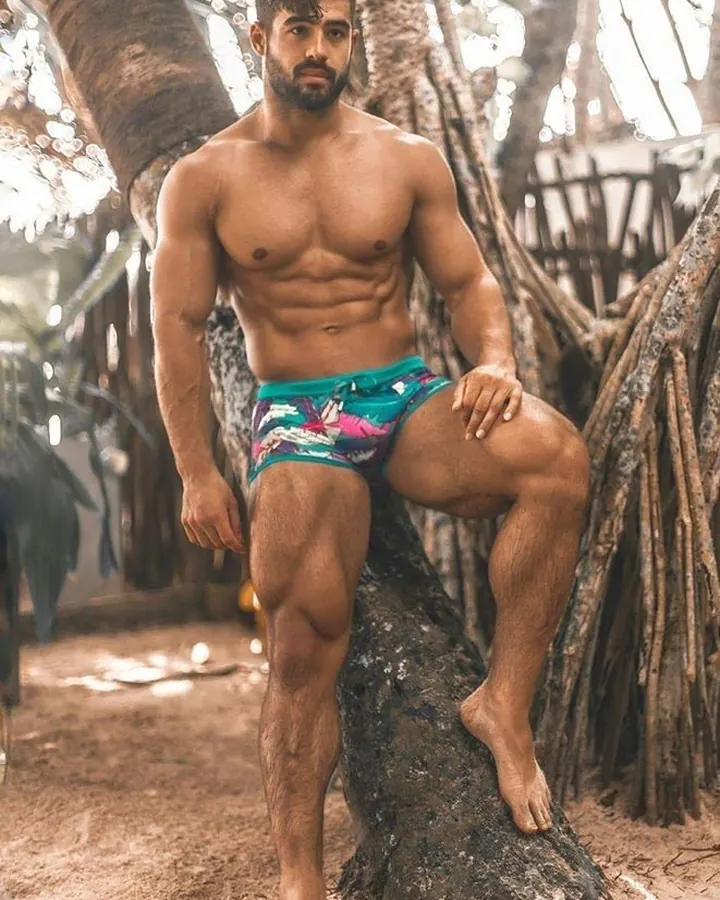 Homme arabe barbu, torse nu, portant un slip Calvin Klein, souriant contre un mur décoré.