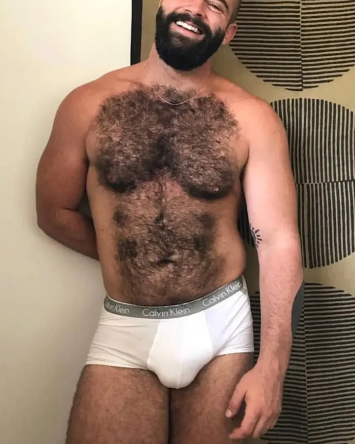 Homme latino barbu, torse nu, portant un slip noir, posant dans une piscine.