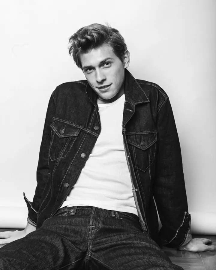 Jeune homme européen aux cheveux blonds, assis sur le sol, vêtu d'une veste en jean et d'un t-shirt blanc.