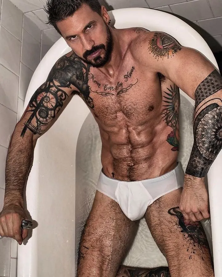 Homme barbu, tatoué, en sous-vêtement blanc, assis dans une baignoire