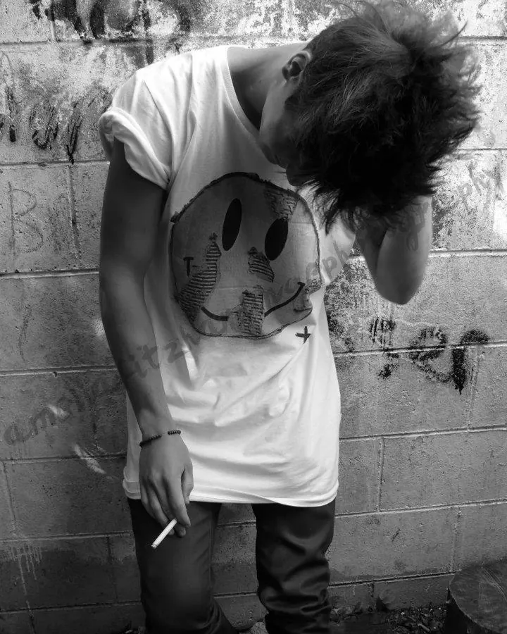 Jeune homme en t-shirt blanc avec un smiley, la tête baissée, fumant une cigarette devant un mur de briques en noir et blanc