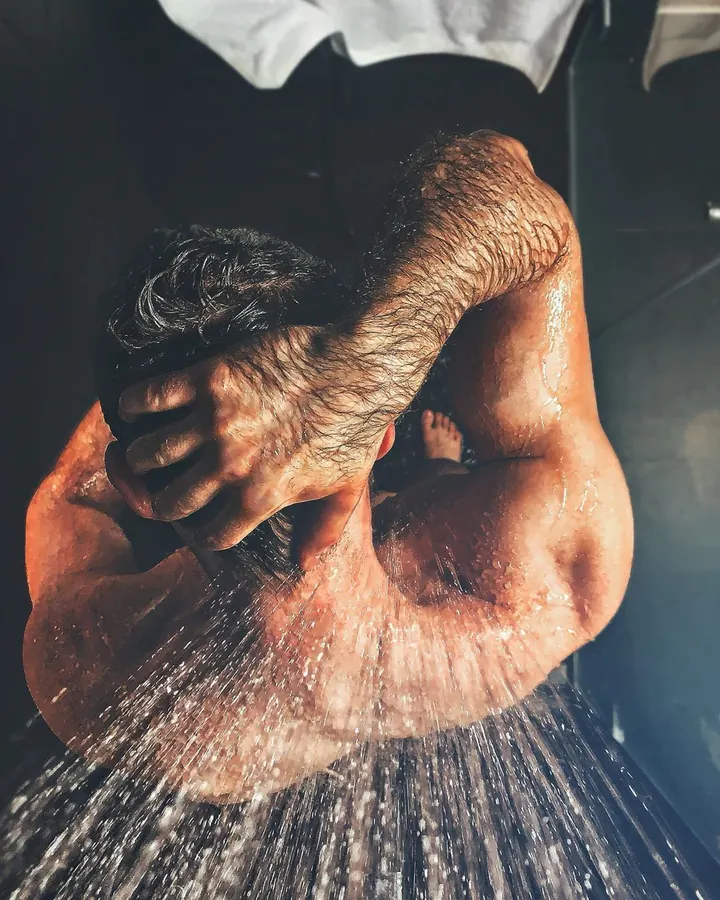 Homme blanc, torse nu, sous la douche, les mains dans les cheveux.