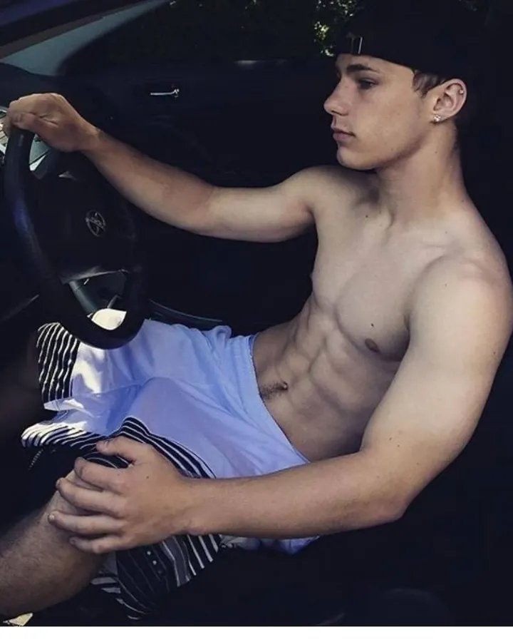 Jeune homme torse nu, en short blanc, assis dans une voiture, regard pensif.