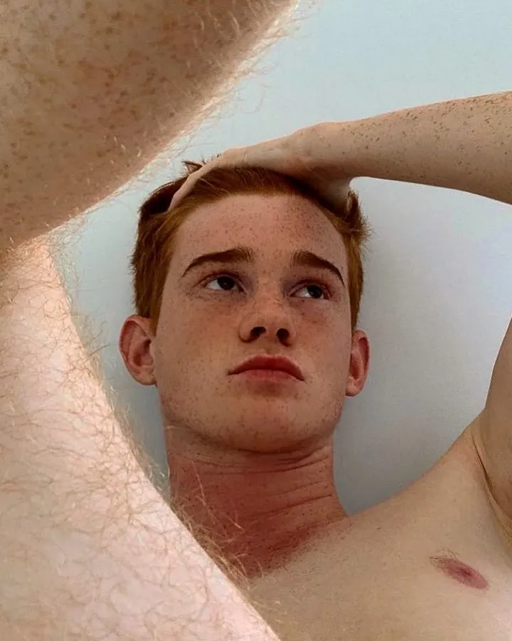 Jeune homme aux cheveux roux, torse nu, main sur la tête, regardant vers le haut, en plan rapproché.