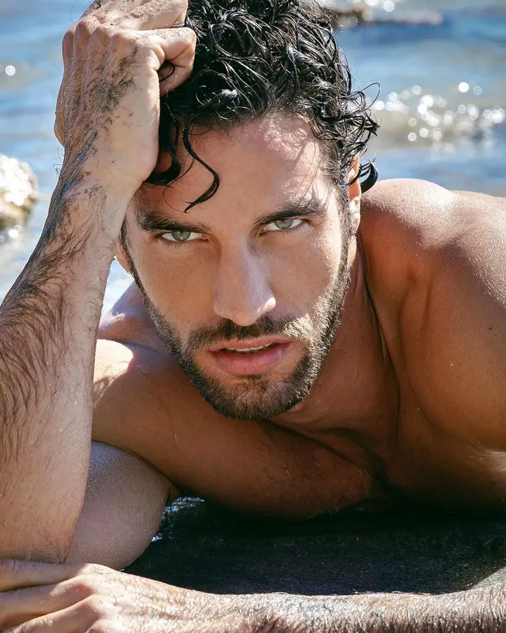 Homme torse nu, barbu, regardant l'appareil, main sur la tête, dans l'eau, soleil brillant.