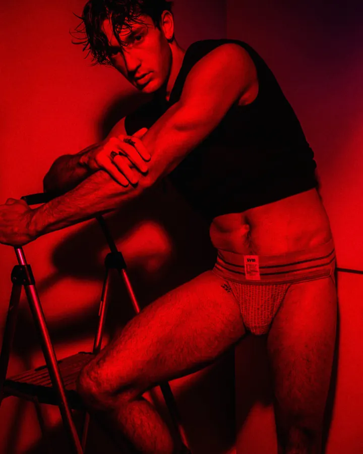 Homme en sous-vêtement rouge, assis sur une échelle, dans une lumière rouge, regardant l'appareil.
