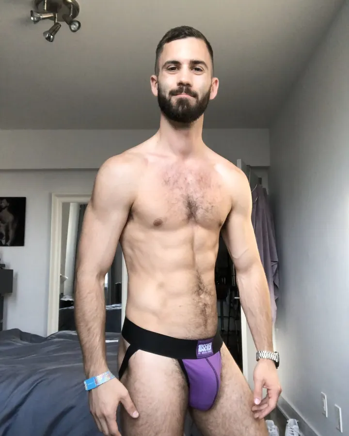 Homme torse nu en caleçon violet, poitrine poilue, barbe fournie, regardant l'appareil dans une chambre.