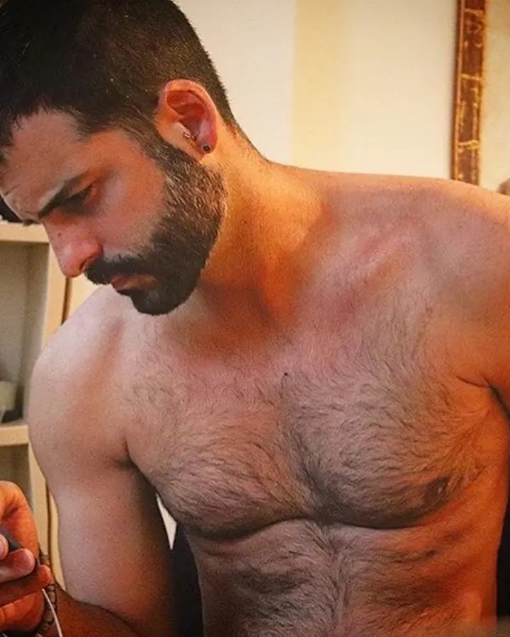 Homme torse nu, barbe fournie, regardant intensément son téléphone portable dans une pièce aux murs clairs.