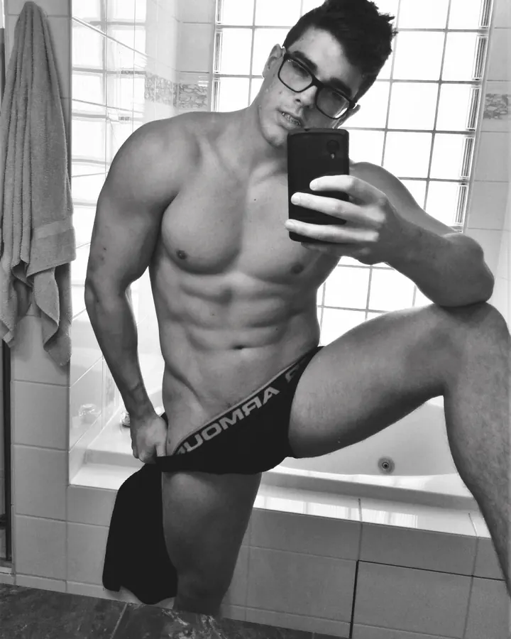 Homme torse nu, portant des lunettes, prenant un selfie dans une salle de bain, tenant son caleçon noir.