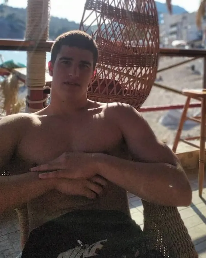 Homme torse nu, assis sur une chaise en osier, mains croisées sur la poitrine, en plein air, soleil éclatant.